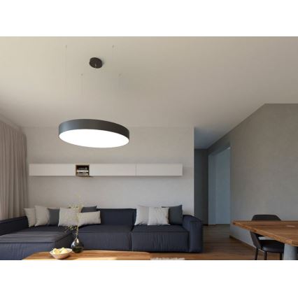 Okrúhle stropné svietidlo svieti v obývacej izbe. Svietidlo visí z nízkeho stropu nad tmavomodrou sedačkou a konferenčným stolíkom. V pozadí je vidieť jedálenský stôl a svetlé steny.