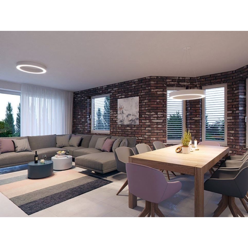 Rohový gauč v obývacej izbe s jedálenským stolom a stoličkami. Steny sú z červených tehál, okná majú žalúzie. Svietidlá sú kruhové, jedno stropné, druhé visiace nad stolom.