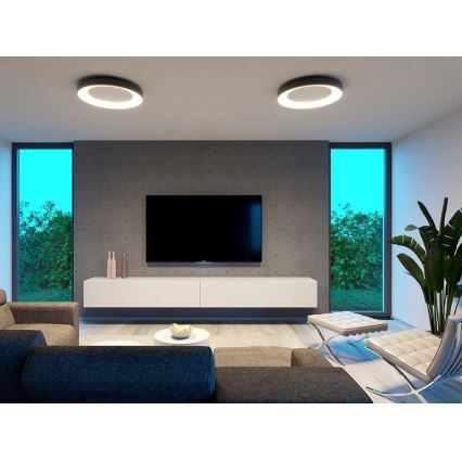 Dve kruhové stropné svietidlá osvetľujú modernú obývačku s televízorom umiestneným na bielej TV zostave. V pozadí sú viditeľné dve okná a betónová stena.