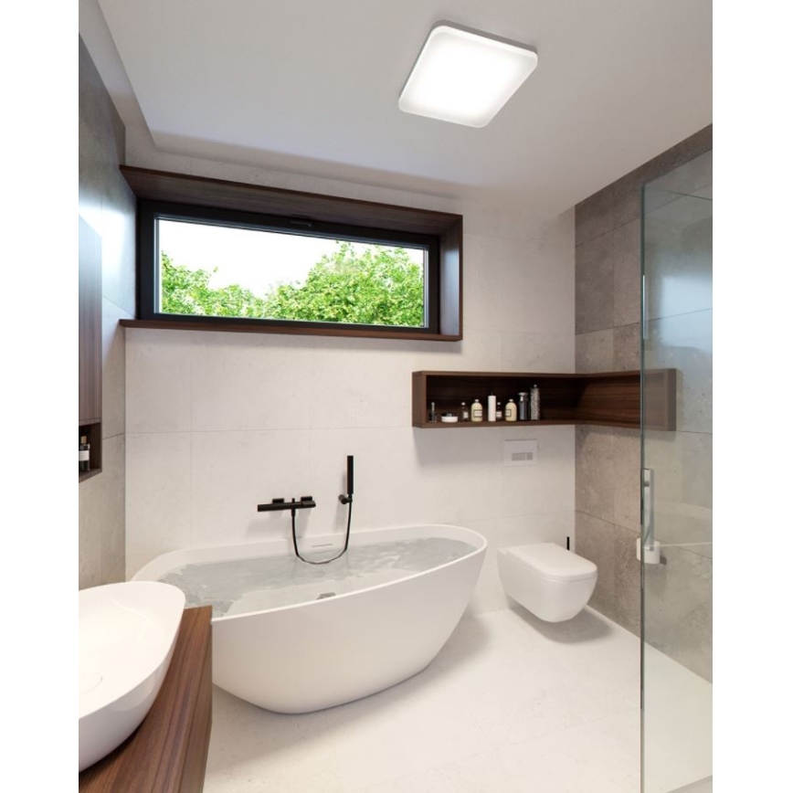 Svietidlo svieti v modernej kúpeľni. V kúpeľni je voľne stojaca vaňa s vodou, umývadlo a závesná toaleta. Okno s výhľadom na zeleň dopĺňa priestor.