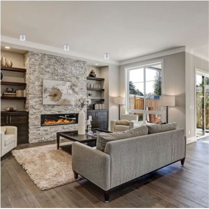 Šedá rozkladacia pohovka stojí v obývacej izbe. V pozadí je krb z kameňa s televízorom nad ním a vstavané police. Podlaha je z dreva.