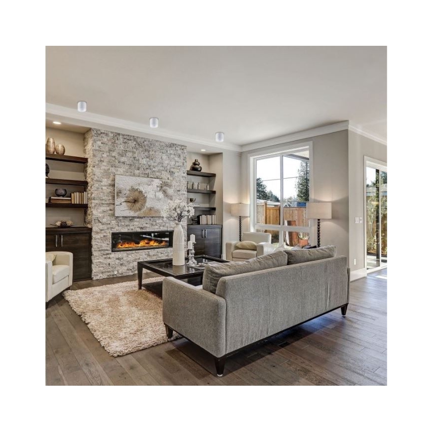 Šedá rozkladacia pohovka stojí v obývacej izbe. V pozadí je krb z kameňa s televízorom nad ním a vstavané police. Podlaha je z dreva.