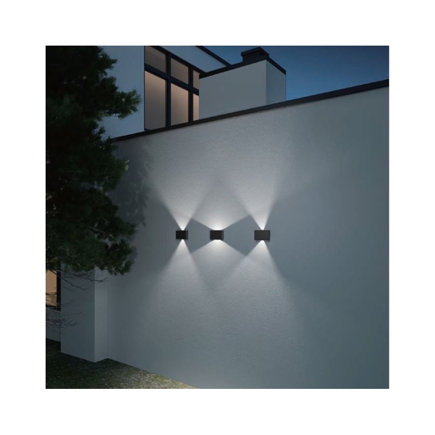 Tri nástenné svietidlá svietia na svetlej vonkajšej stene domu. Svietidlá sú štvorcové, čierne a sú umiestnené vedľa seba. V pozadí je viditeľná časť domu a strom.