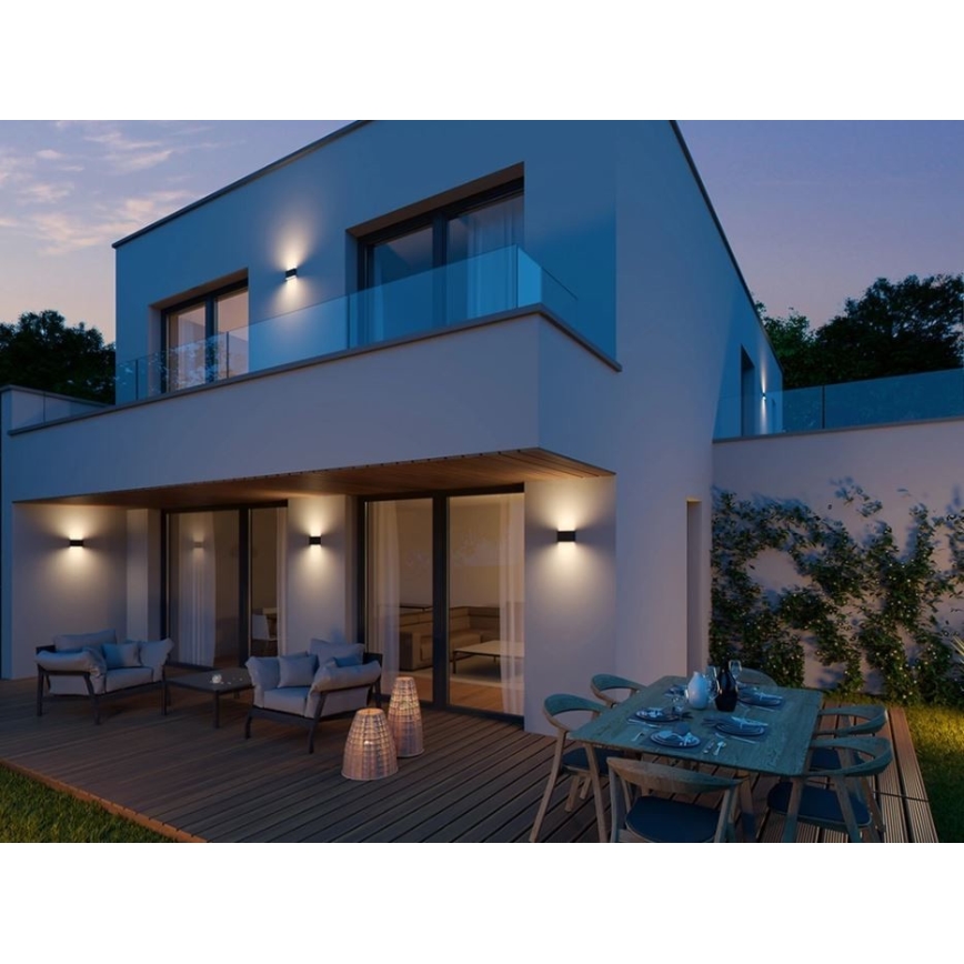 Moderná vila s nástennými svietidlami svieti večer. Svietidlá osvetľujú terasu s nábytkom a jedálenským stolom. Okolím je zeleň.