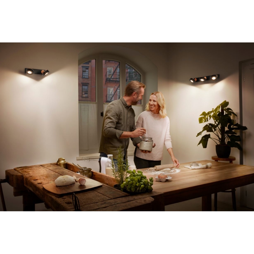 Dvojica pripravuje cesto na drevenom stole v kuchyni osvetlenej dvoma nástennými svietidlami s tromi bodovými svetlami. Na stole sú viditeľné ingrediencie ako múka, vajíčka a bylinky.