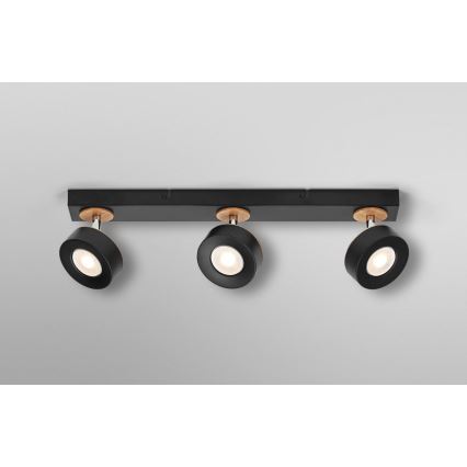 Trojité stropné svietidlo s čiernymi reflektormi a drevenými detailmi osvetľuje priestor. Reflektor je otočný, pripevnený k čiernej lište. Lišta je upevnená na svetlom sivom strope.