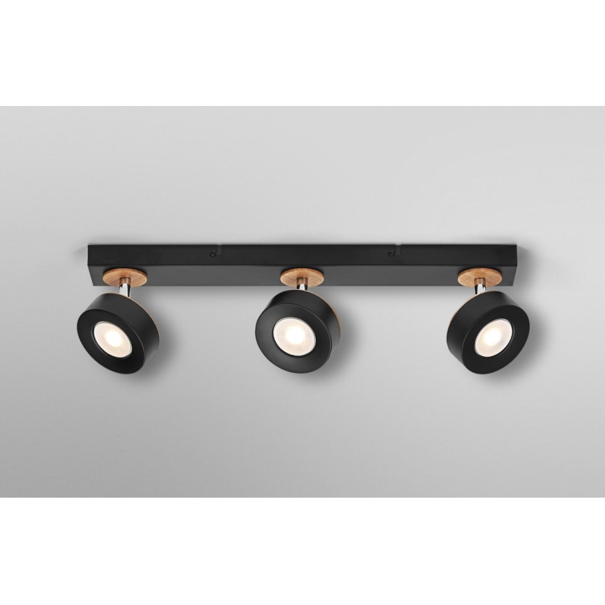 Trojité stropné svietidlo s čiernymi reflektormi a drevenými detailmi osvetľuje priestor. Reflektor je otočný, pripevnený k čiernej lište. Lišta je upevnená na svetlom sivom strope.