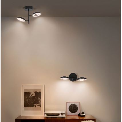 Dve trojramenné svietidlá svietia v interiéri. Jedno je pripevnené na stene, druhé na strope. Svietidlá sú umiestnené v miestnosti s nábytkom, gramofónom a obrazmi.