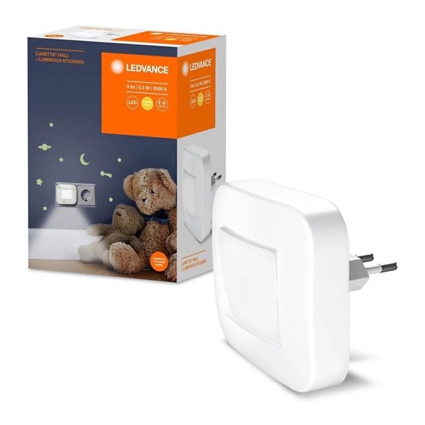 Biela LED nočná lampa svieti v detskej izbe. Lampa je zapojená do zásuvky. Na krabici je uvedené: LUNETTA HALL + LUMINOUS STICKERS, LEDVANCE, 3 lm 0,2 W 3000 K.