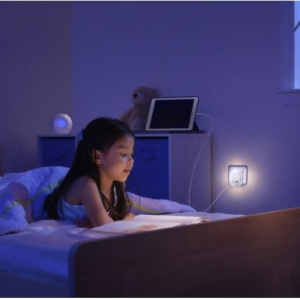 Dievča v detskej izbe leží v posteli a číta knihu pri svetle nočnej lampy zapojenej do zásuvky. V pozadí je plyšový medveď a tablet.