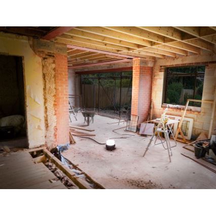 Svietidlo svieti v interiéri rozostavaného domu. V miestnosti je rozsiahly stavebný neporiadok: stavebné materiály, náradie, otvorené murivo a odstránené časti podlahy. Vidieť drevené trámy stropu, tehlové steny a otvorený priestor do záhrady.