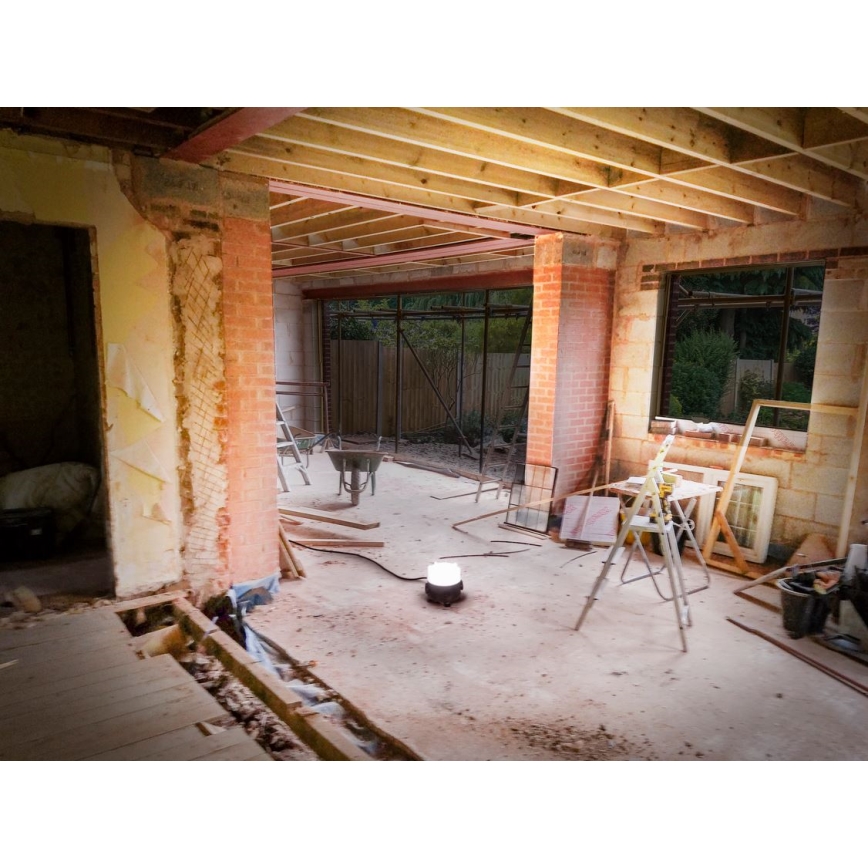 Svietidlo svieti v interiéri rozostavaného domu. V miestnosti je rozsiahly stavebný neporiadok: stavebné materiály, náradie, otvorené murivo a odstránené časti podlahy. Vidieť drevené trámy stropu, tehlové steny a otvorený priestor do záhrady.