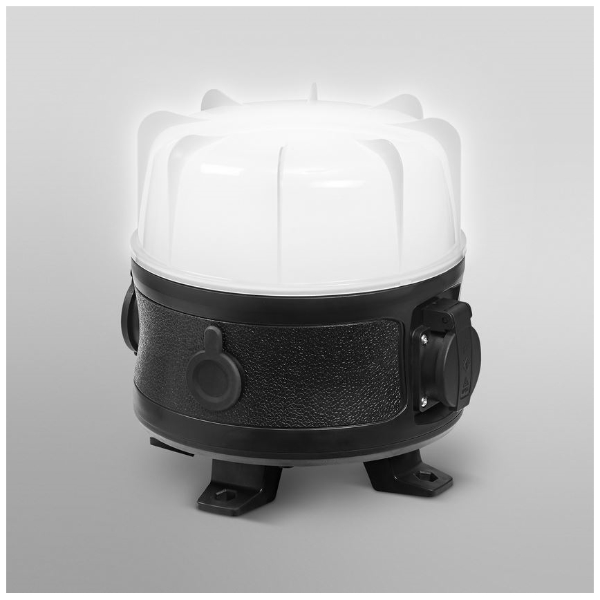 Prenositelná lampa svieti. Je umiestnená na neutrálnom pozadí. Lampa má biely vrchný diel a čierny spodný diel s vypínačom a elektrickou zásuvkou. Má tiež štyri nožičky.