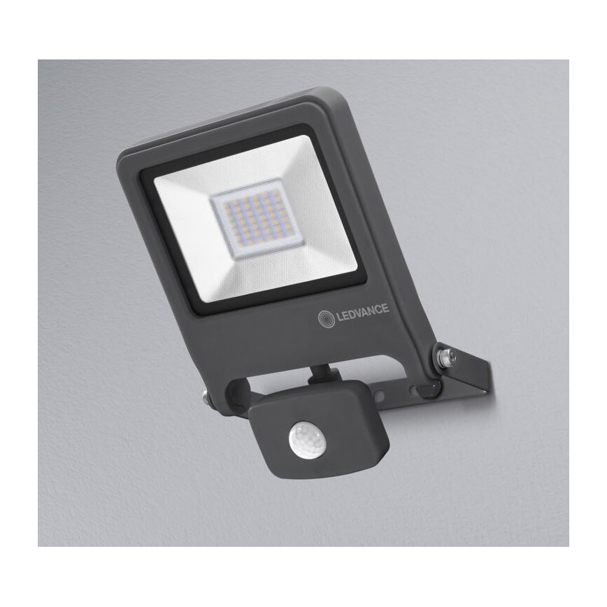LED reflektor s pohybovým senzorom je pripevnený na stenu. Svietidlo je sivé a obsahuje LED diódy. Na obrázku je viditeľné logo LEDVANCE.