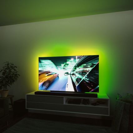 Televízor zobrazuje zábery pretekov áut. Je umiestnený na televíznom stolíku v miestnosti s tlmeným zeleným podsvietením. Na stolíku sú ďalšie zariadenia.
