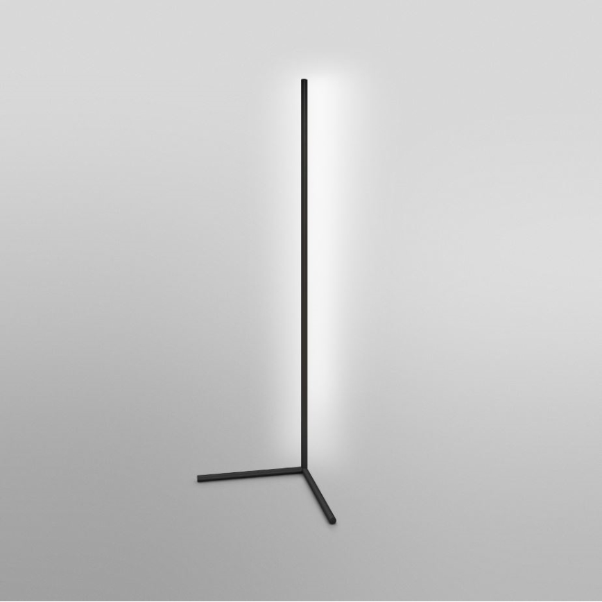 Stojaca lampa svieti. Má vysoký čierny stĺpik a trojnohý podstavec. Nachádza sa na svetlej podlahe.