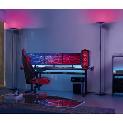 Herná stolička s červenými a čiernymi prvkami stojí pri hernom stole s troma monitormi a počítačovou vežou. Dve vysoké stojace lampy s farebným osvetlením osvetľujú miestnosť. V pozadí je ďalší nábytok.