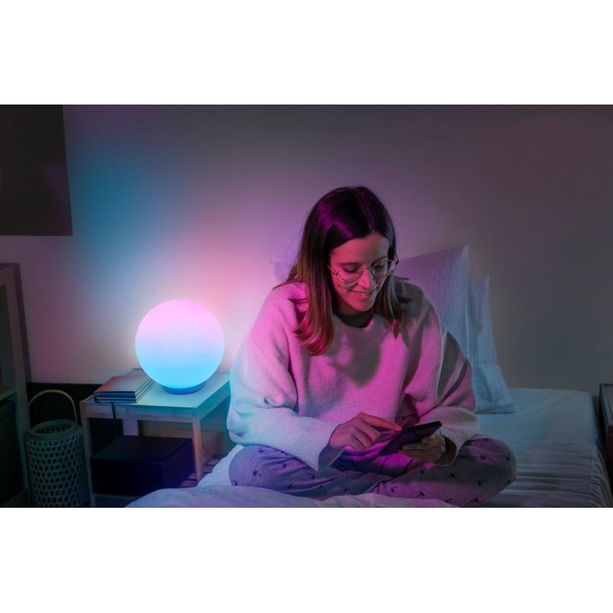 Žena sedí na posteli v osvetlenej miestnosti a používa mobilný telefón. Pri posteli je guľatá lampa s prechodom farieb od modrej po ružovú.