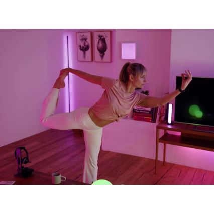 Žena vykonáva jógovú pozíciu v svetlej miestnosti s drevenou podlahou, s farebným osvetlením a televízorom v pozadí.