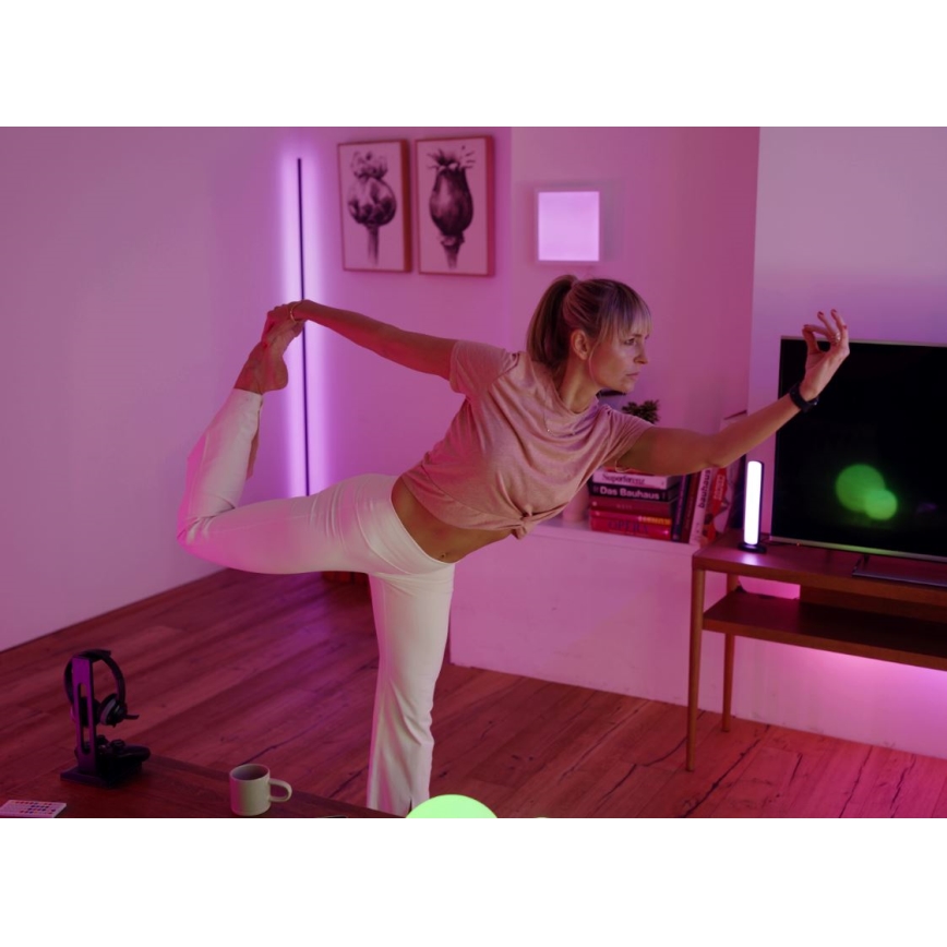 Žena v bielych nohaviciach a svetloružovom tielku vykonáva jogové cvičenie v miestnosti s drevenou podlahou a farebným osvetlením. V pozadí je televízor a nábytok.