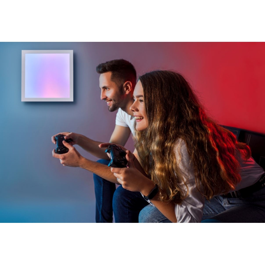 Pár hrá video hru s ovládačmi, sediac na pohovke v miestnosti s farebným podsvietením. Na stene je umiestnený štvorcový svetelný panel s premenlivými farbami.