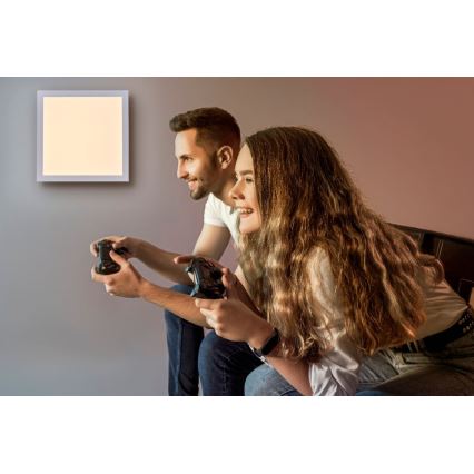 Pár hrá hru na konzolu s ovládačmi v ruke. Sedia na posteli pred štvorcovou nástennou lampou s teplým svetlom. Izba má neutrálnu farbu stien.