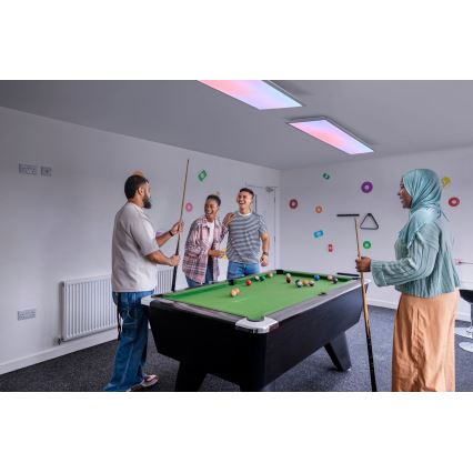 Štyria mladí ľudia hrajú biliard v miestnosti s bielymi stenami a farebnými dekoráciami na stenách. Dvaja držia tága a dvaja sa usmievajú pri stole.