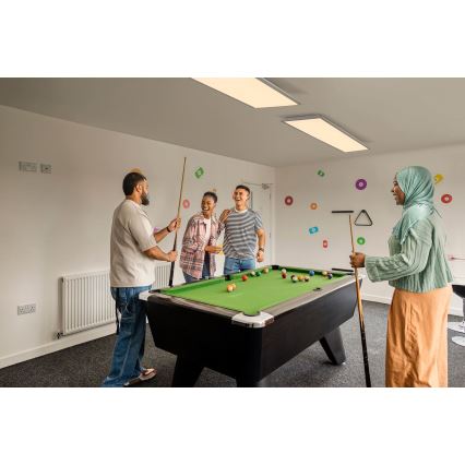 Štyria ľudia hrajú biliard v miestnosti s bielymi stenami a farebnými kruhmi na stenách. Jeden muž drží biliardovú taku. Ostatní sa prizerajú a smejú.