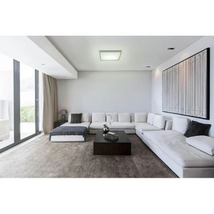 Svietidlo svieti v modernej obývacej izbe. Svetlo osvetľuje bielu rohovú sedačku a konferenčný stolík. Izba má veľké okná a moderný obraz na stene.