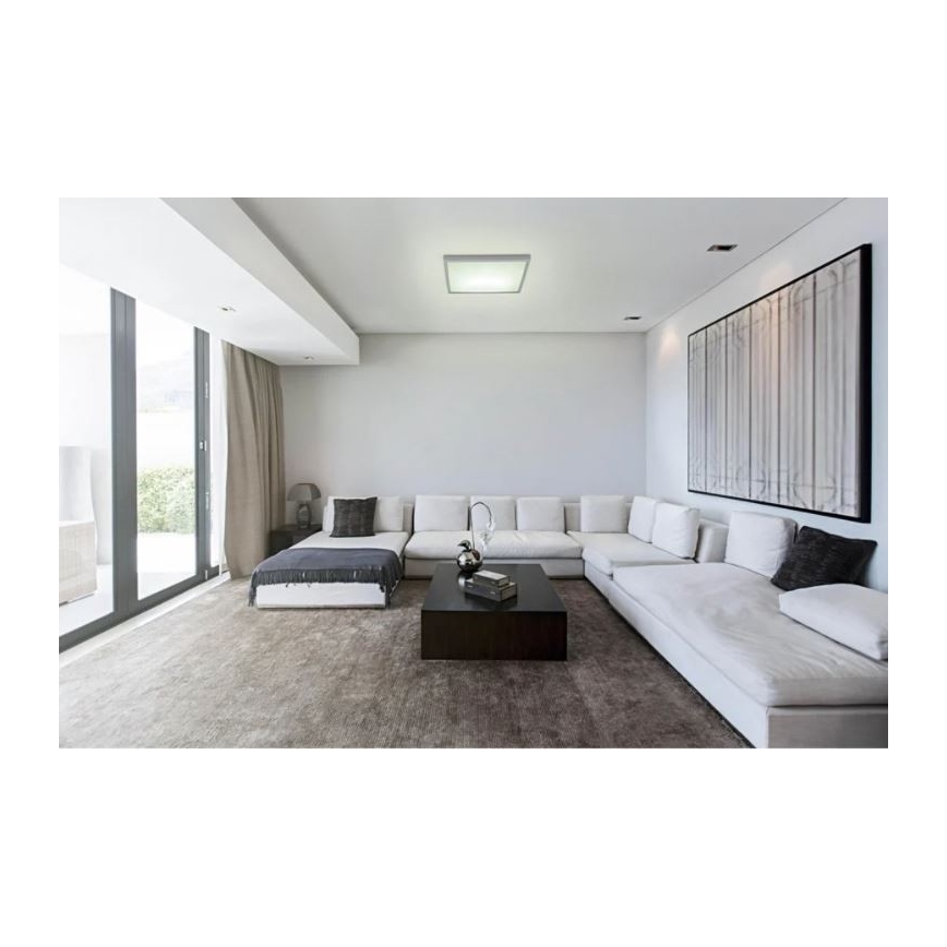 Svietidlo svieti v modernej obývacej izbe. Svetlo osvetľuje bielu rohovú sedačku a konferenčný stolík. Izba má veľké okná a moderný obraz na stene.