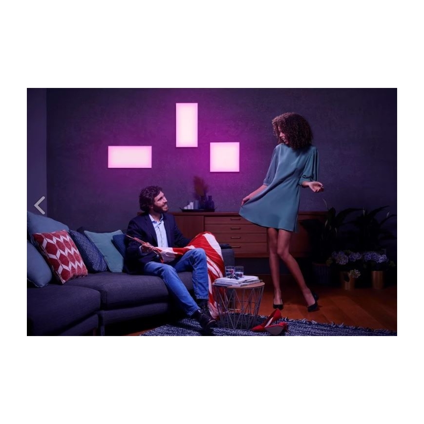 Muž sedí na pohovke, číta knihu. Žena tancuje v šatách. Na stene sú tri štvorcové svetlá s fialovým svetlom. Prostredie tvorí obývacia izba.