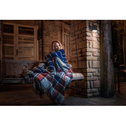 Žena sedí zabalená v plede na záhradnej hojdačke. Na stene vedľa nej svieti nástenné vonkajšie svietidlo. Prostredie je nočné, na kamennom terase pri dome.