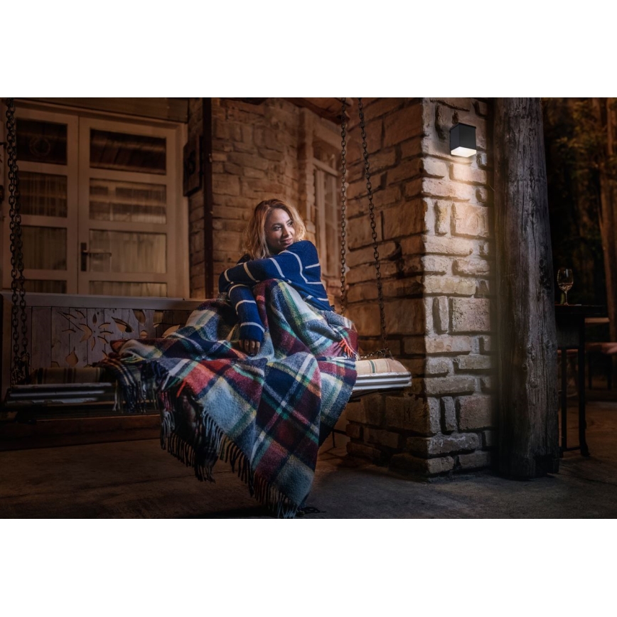 Žena sedí zabalená v plede na záhradnej hojdačke. Na stene vedľa nej svieti nástenné vonkajšie svietidlo. Prostredie je nočné, na kamennom terase pri dome.