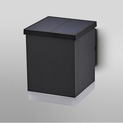 Nástená solárna lampa svieti. Je štvorcového tvaru, tmavosivej farby, s horným solárnym panelom a spodným svetelným zdrojom. Pripevnená je na stenu.