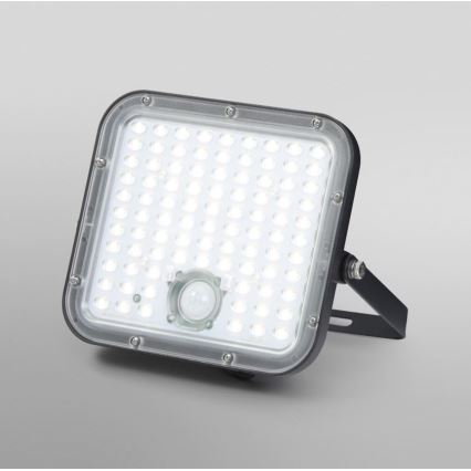 LED reflektor s pohybovým senzorom svieti. Je štvorcového tvaru, má čierny rám a je umiestnený na svetlomodrom pozadí.