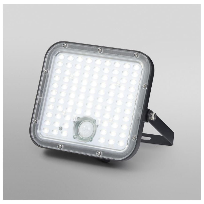 LED reflektor s pohybovým senzorom svieti. Je štvorcového tvaru, má čierny rám a je umiestnený na svetlomodrom pozadí.