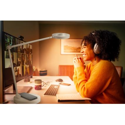 Stolová lampa s bezdrôtovou nabíjačkou osvetľuje ženu počas online konferencie. Žena v slúchadlách sedí pri stole s počítačom, klávesnicou a poznámkovým blokom. Svetlo lampy je teplé.