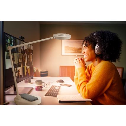 Žena s náhlavnou súpravou sa usmieva počas online konferencie na monitore. Na stole je lampa s bezdrôtovou nabíjačkou pre mobilný telefón, klávesnica a zápisník.