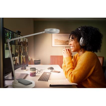 Stolná lampa s bezdrôtovou nabíjačkou osvetľuje pracovisko ženy počas online konferencie. Žena s náhlavnou súpravou sedí pri počítači s rozsiahlym videokonferenčným oknom. Na stole sa nachádza klávesnica, telefón a zápisník.