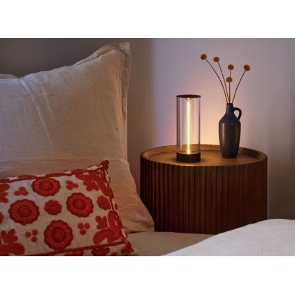 Svietidlo svieti na nočnom stolíku v spálni. Stojí na tmavom drevenom stolíku vedľa vázy so suchými kvetmi. V pozadí je posteľ s vankúšmi.