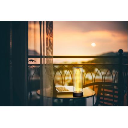 Stolná lampa svieti na balkóne pri západe slnka. Lampa je vysoká, valcového tvaru, s teplým žltým svetlom vnútri. Stojí na okrúhlom stole vedľa notebooku.