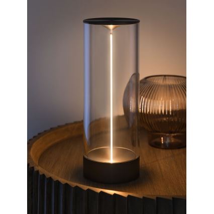 Stolná lampa svieti. Svetlo vychádza z úzkeho, vertikálne umiestneného svetelného zdroja v sklenenom valci s tmavou kovovou základňou. Lampa stojí na drevenom podnose.