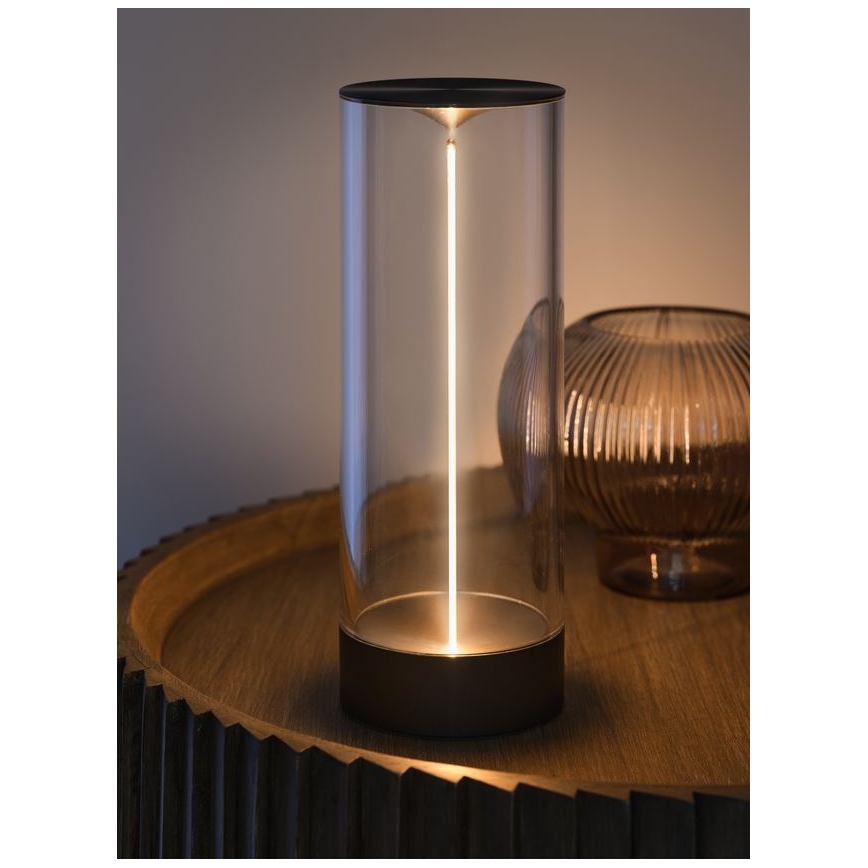 Stolná lampa svieti. Svetlo vychádza z úzkeho, vertikálne umiestneného svetelného zdroja v sklenenom valci s tmavou kovovou základňou. Lampa stojí na drevenom podnose.