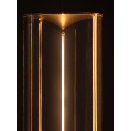 Svetlo svieti v priehľadnom valcovom sklenenom obale s kovovými prvkami. Svetlo je umiestnené vertikálne v strede. Pozadie je tmavé.