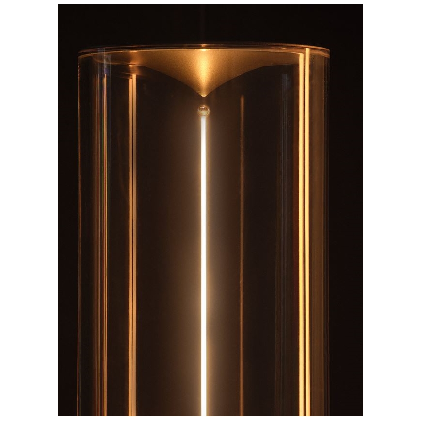 Svetlo svieti v priehľadnom valcovom sklenenom obale s kovovými prvkami. Svetlo je umiestnené vertikálne v strede. Pozadie je tmavé.