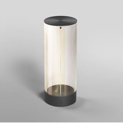 Svietidlo svieti. Sklenený valec s tmavosivými vrchným a spodným krytom obsahuje tenkú, vertikálne orientovanú svetelnú tyčinku. Svietidlo stojí na svetlošedej ploche.