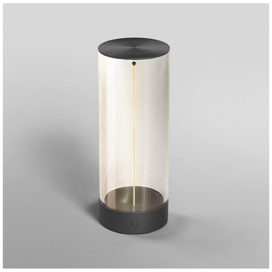 Svietidlo svieti. Sklenený valec s tmavosivými vrchným a spodným krytom obsahuje tenkú, vertikálne orientovanú svetelnú tyčinku. Svietidlo stojí na svetlošedej ploche.