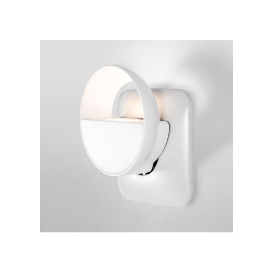 Biela nočná lampa v tvare polkruhu svieti. Zariadenie je zapojené do bielej elektrickej zásuvky na stene.