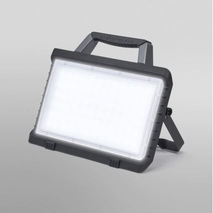 Prenositelná LED pracovná lampa svieti. Má robustný tmavosivý plastový kryt, rúčkový úchyt a výklopný stojan. Lampa je umiestnená na svetlom pozadí.