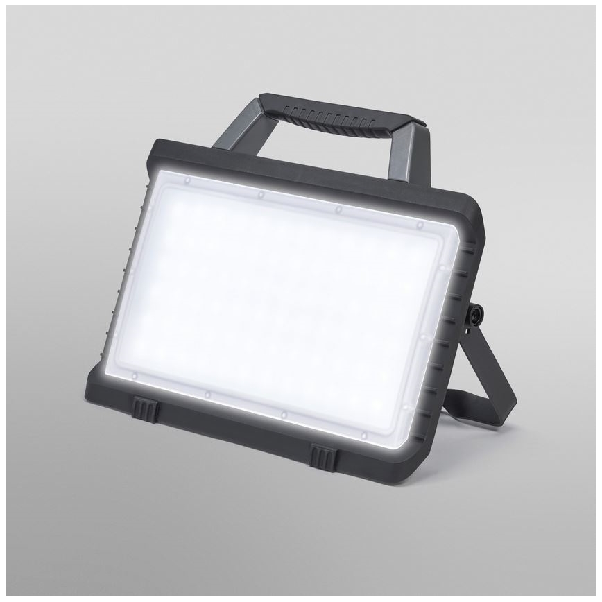 Prenositelná LED pracovná lampa svieti. Má robustný tmavosivý plastový kryt, rúčkový úchyt a výklopný stojan. Lampa je umiestnená na svetlom pozadí.